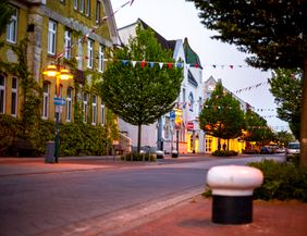 Eine ruhige, mit Bäumen gesäumte Straße in einer Stadt bei Dämmerung, geschmückt mit bunten Wimpeln und beleuchtet von Straßenlaternen.