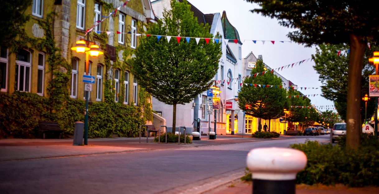 Eine ruhige, mit Bäumen gesäumte Straße in einer Stadt bei Dämmerung, geschmückt mit bunten Wimpeln und beleuchtet von Straßenlaternen.